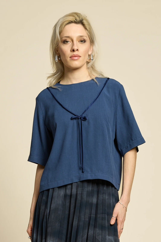 A woman wearing the Lea Top by Cokluch in Denim, a loose top with short dropped sleeves, a round neck, and an attached V-shaped ribbon with a bow and ties. She is wearing it with black skirt and standing in front of a beige wall. 