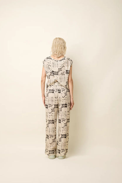 Back view of a woman wearing the Bayang Shirt by Cokluch in Natural Ink, featuring a geometric print, round neck, short extended sleeves, and a cross-over bottom hem at the back. She is wearing it with matching pants and standing in front of a beige background. 