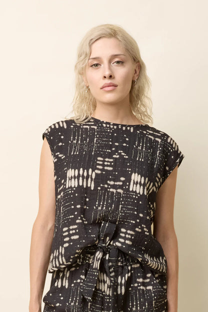 A woman wearing the Bayang Shirt by Cokluch in Black Ink, featuring a geometric print, round neck, short extended sleeves, and a cross-over bottom hem at the back. She is wearing it with matching bottoms and standing in front of a beige background. 