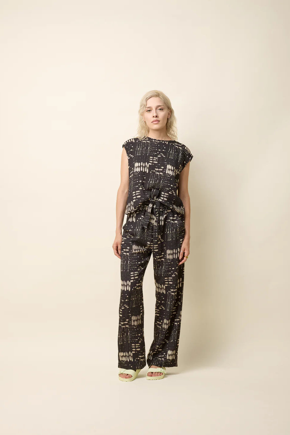  A woman wearing the Bayang Shirt by Cokluch in Black Ink, featuring a geometric print, a round neck, short extended sleeves, and a cross-over bottom hem at the back. She is wearing it with matching pants and standing in front of a beige background.