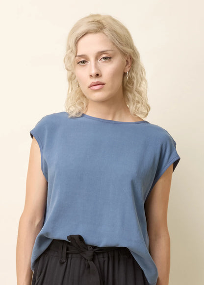 A woman wearing the Bayang Shirt by Cokluch in Ocean, featuring a  round neck, short extended sleeves, and a cross-over bottom hem at the back. She is wearing it with black pants and is standing in front of a beige background. 