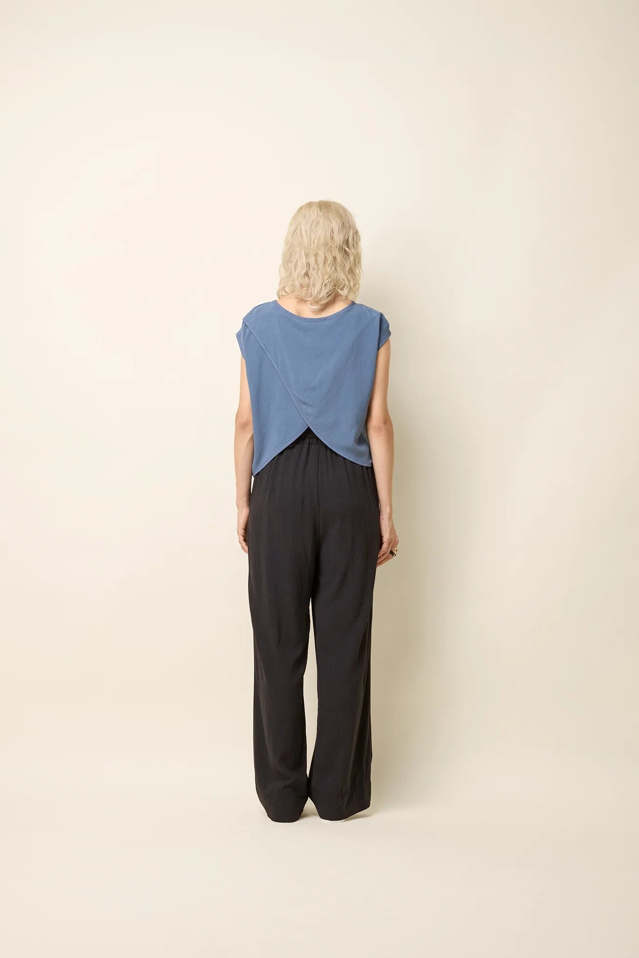Back view of a woman wearing the Bayang Shirt by Cokluch in Ocean, featuring a  round neck, short extended sleeves, and a cross-over bottom hem at the back. She is wearing it with black pants and is standing in front of a beige background. 