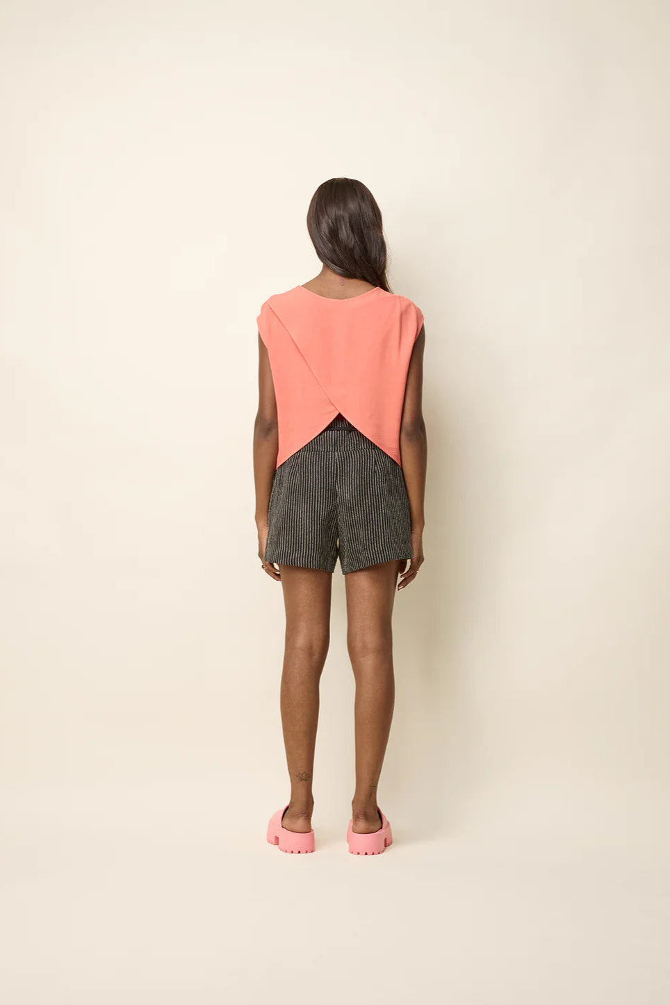 Back view of a woman wearing the Bayang Shirt by Cokluch in Papaya featuring a round neck, short extended sleeves, and a cross-over bottom hem at the back. She is wearing it with striped shorts and standing in front of a beige background.