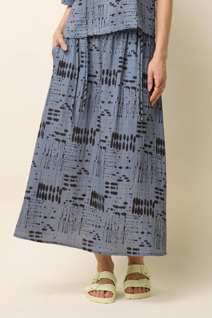 Waist-down view of a woman wearing the Susan Skirt by Cokluch in Blue Ink, an ankle-length skirt in a geometric print with an elastic waist with double drawstrings, a side slit, and side pockets. She is standing in front of a beige background. 