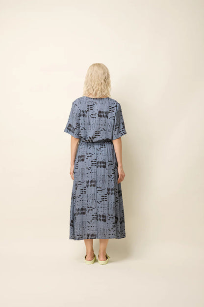 Back view of a woman wearing the Susan Skirt by Cokluch in Blue Ink, an ankle-length skirt in a geometric print with an elastic waist with double drawstrings, a side slit, and side pockets. She is wearing a matching top and is standing in front of a beige background. 