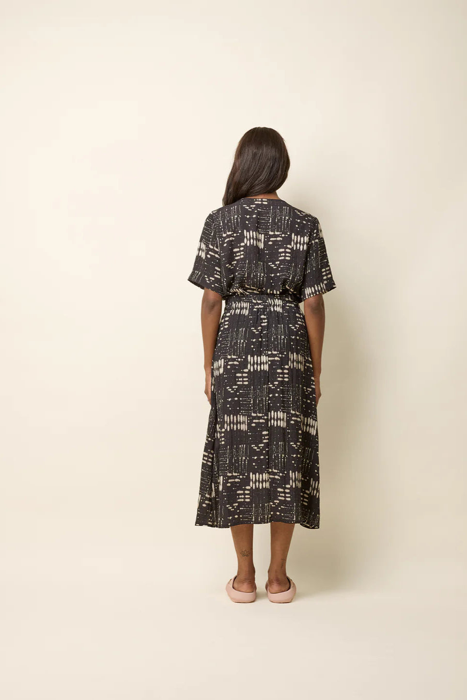 Back view of a woman wearing the Susan Skirt by Cokluch in Black Ink, an ankle-length skirt in a geometric print with an elastic waist with double drawstrings, a side slit, and side pockets. She is wearing a matching top and is standing in front of a beige background. 