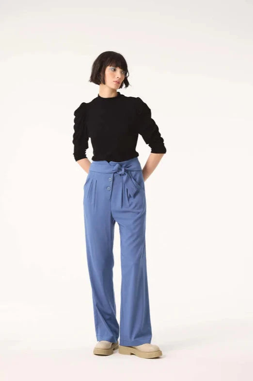 A woman wearing the Nenuphar Top by Cokluch in Black Bamboo, featuring a high collar and 3/4 length puffed sleeves, with light blue pants. She standing in front of a white background. 