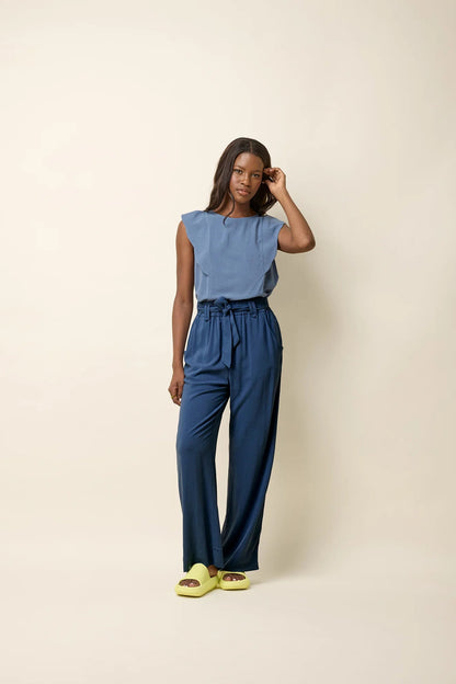 A woman wearing the Yael Pants by Cokluch in Ocean, slightly flared pants with an elastic waist, tie belt with pointed belt loops, and side pockets. She is wearing them with the Neema top in Ocean and standing in front of a beige background. 