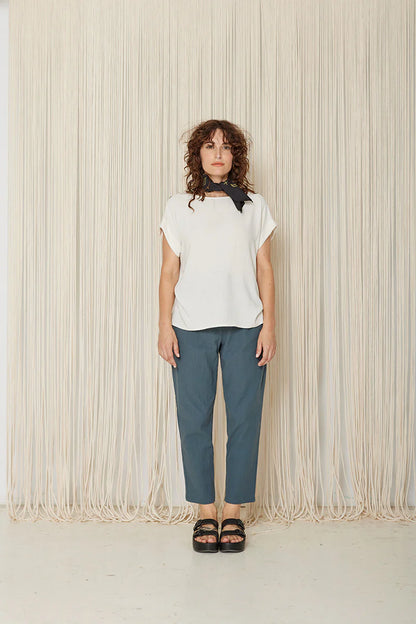 A woman wearing the Yasmina Pants from Cokluch in Steel, with a high elastic waist with a ring belt, and tapered, ankle-length legs. She is wearing them with a white top and standing in front of a beige curtain. 