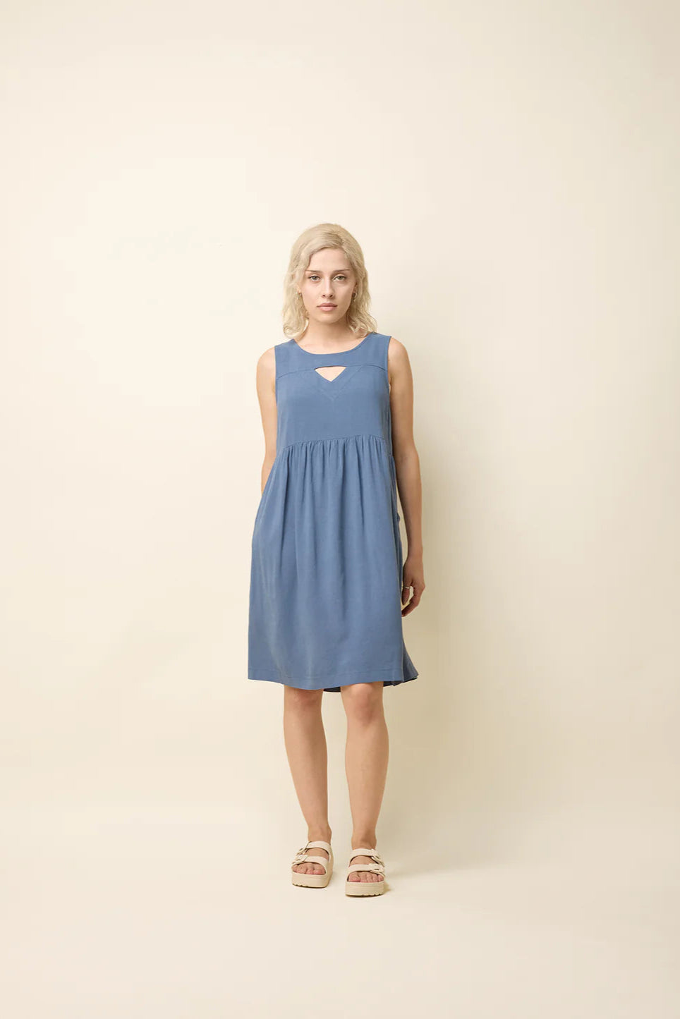 A woman wearing the Esi Dress by Cokluch in Ocean, a sleeveless above-the-knee dress with a round neck, a triangle cutout below the neckline, a empire waist, and a loose fit. She's standing in front of a beige background. 