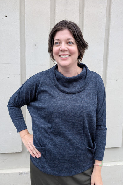 A woman wearing the Nash Sweater by Compli K in Navy, with a cowl neck, long extended sleeves, angled vertical seams on the front, and front patch pockets. She is standing outside in front of a white wall. 