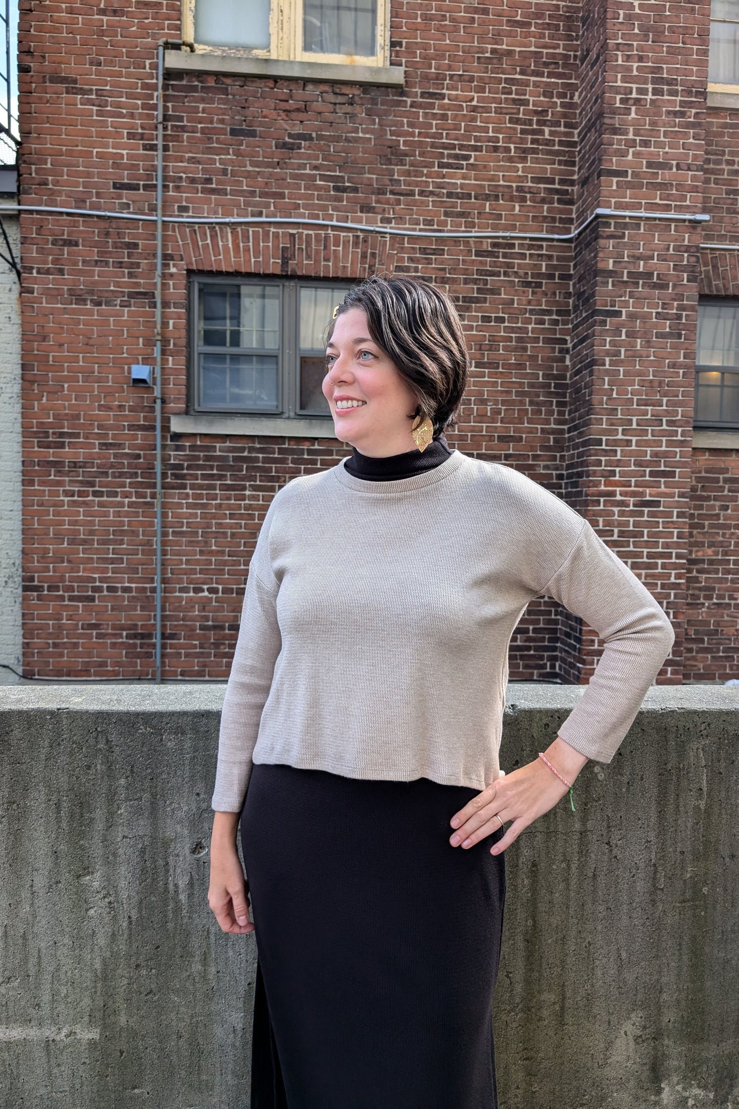 Side view of a woman wearing the Natasha Dress by Compli K in Black, a sleeveless midi-length dress with a mock turtleneck and side slits, under the Nerissa Knit Top in Beige, standing outside in front of a brick wall. 