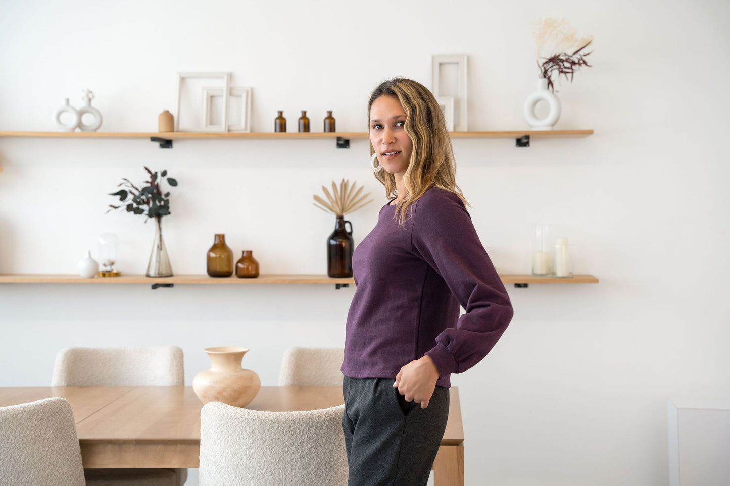 A woman wearing the Cosmopolitain Sweater by Cherry Bobin in Mauve, with a rounded neck and long sleeves with a puff detail. She is wearing it with black pants and standing in a room with shelves. 