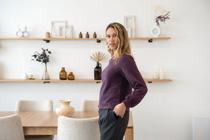 A woman wearing the Cosmopolitain Sweater by Cherry Bobin in Mauve, with a rounded neck and long sleeves with a puff detail. She is wearing it with black pants and standing in a room with shelves. 