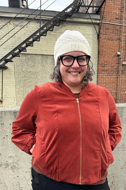 A woman wearing the Beret by Charlevoix Pure Laine in Natural with the Redrawn Zip Jacket by Eve Lavoie in Terracotta Corduroy. She is standing outside. 