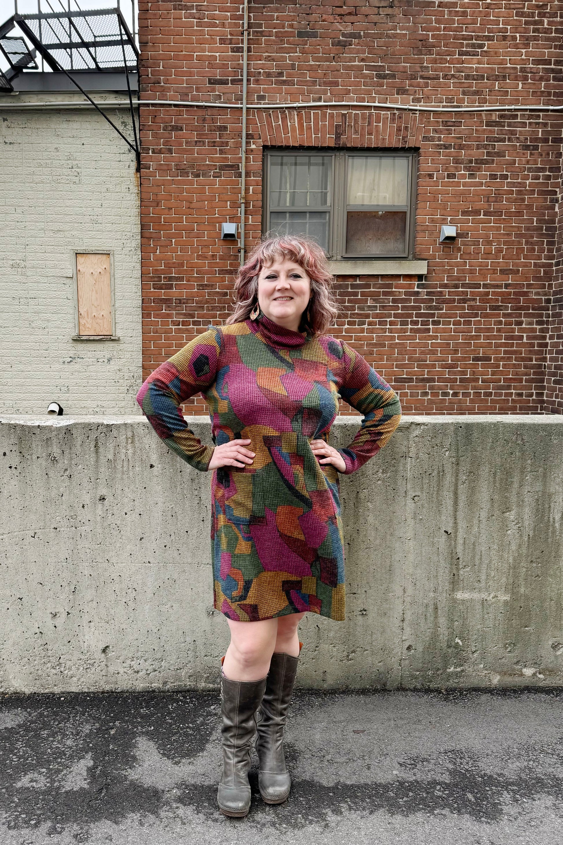 A woman wearing the Damon Dress by Pure Essence in Berry/Mustard, featuring a cowl neck, long sleeves, patch pockets, and a knee length hem. She's standing outside in front of a brick building. 