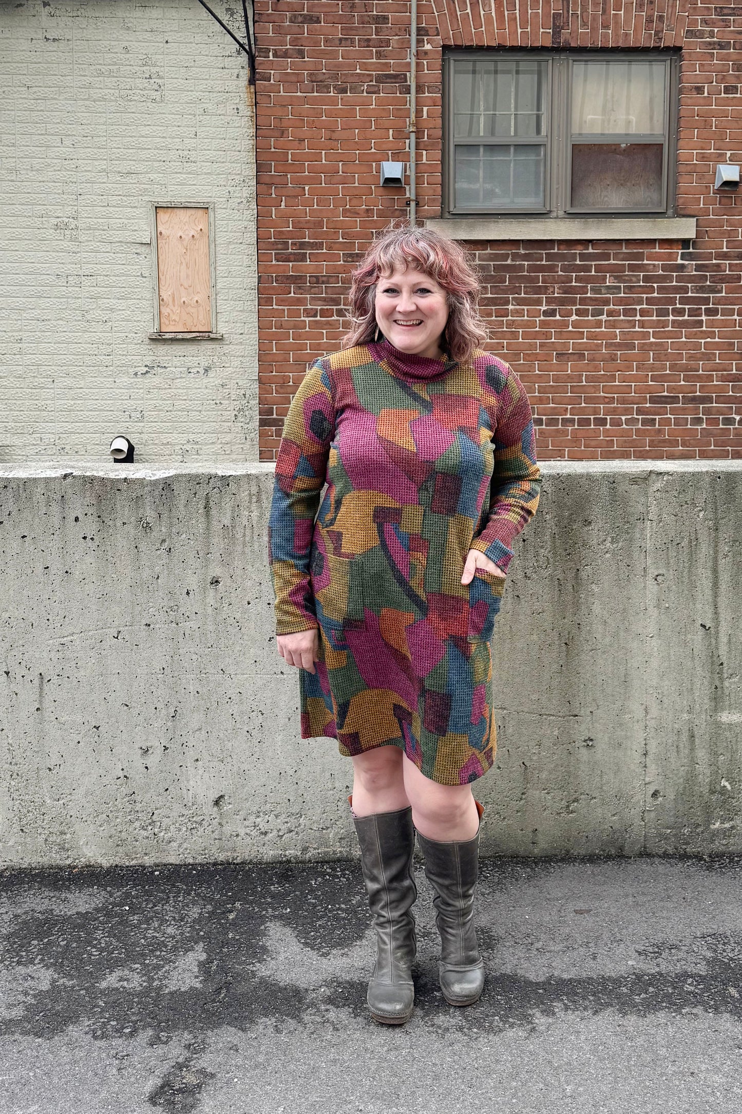 A woman wearing the Damon Dress by Pure Essence in Berry/Mustard, featuring a cowl neck, long sleeves, patch pockets, and a knee length hem. She's standing outside in front of a brick building. 