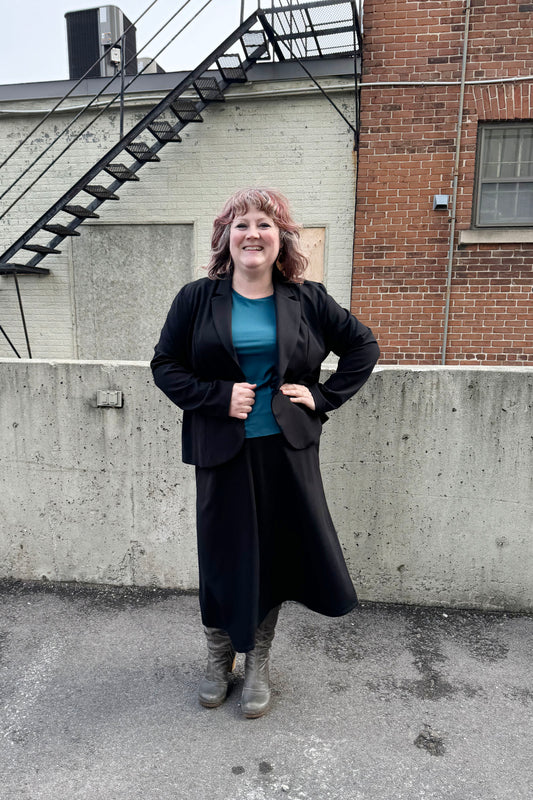 A woman wearing the Famke Jacket by Pure Essence in Black, a single breasted blazer with a single button closure. She is wearing it with a turquoise top and a black skirt and standing in front of a brick wall.