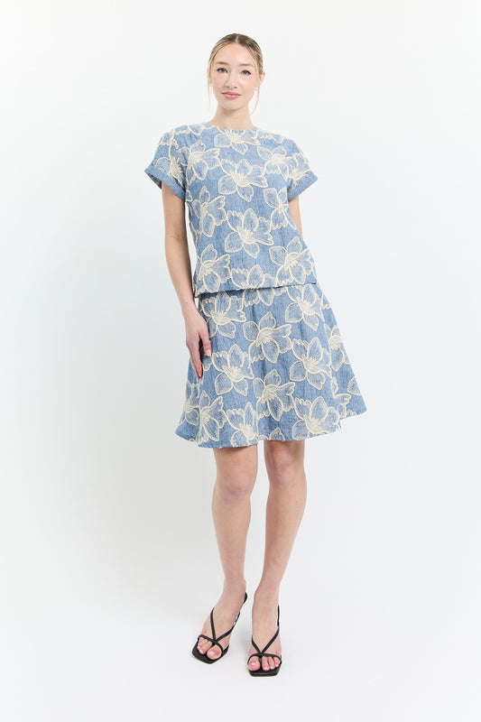 A woman wearing the Sun Top by by Dinh Ba in Blue with white embroidered flowers, with a round neck and short sleeves. She is wearing it with the matching Joy Skirt and standing in front of a white background. 