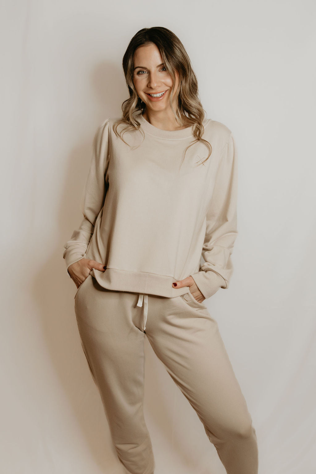 A woman wearing the Tasia Sweatshirt by Betty x Bow in Crema, an oversized crew neck with subtly puffed shoulders, ribbing at the cuffs, hem and collar, and a monochromatic embroidered bow logo. She is wearing it with cream pants and standing in front of a white background. 