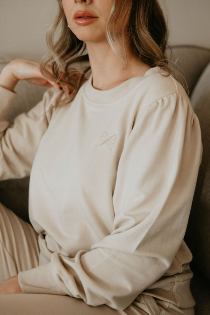 Close up view of a woman wearing the Tasia Sweatshirt by Betty x Bow in Crema, an oversized crew neck with subtly puffed shoulders, ribbing at the cuffs, hem and collar, and a monochromatic embroidered bow logo. d. 