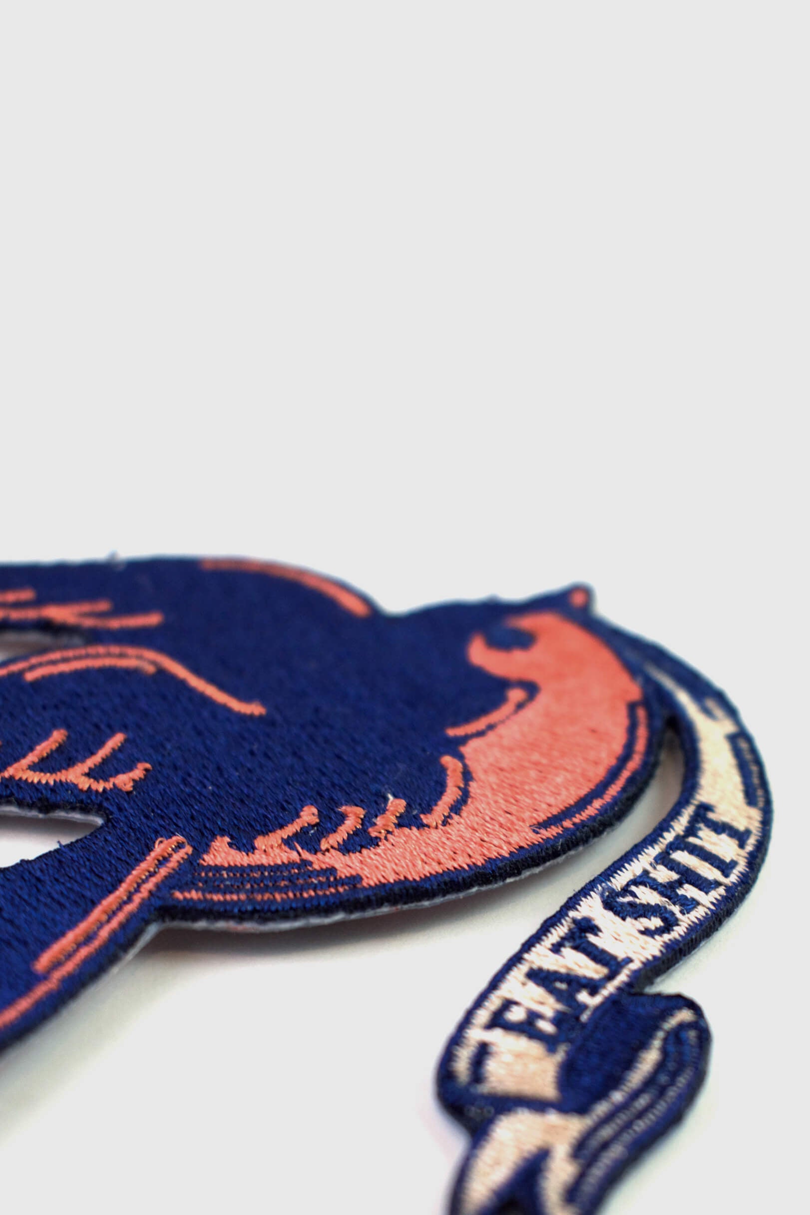 A vintage-style embroidered patch from Badaboöm Studio, 
featuring a flying bird in navy blue and orange, with a banner reading 'EAT SHIT' below, shown on a plain light grey background.