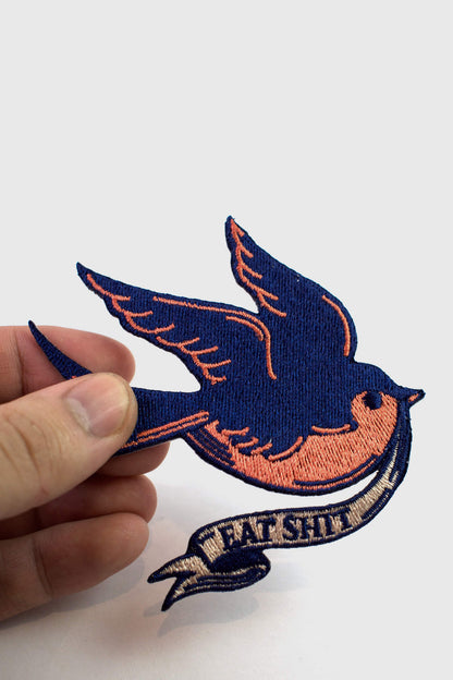 A vintage-style embroidered patch from Badaboöm Studio, 
featuring a flying bird in navy blue and orange, with a banner reading 'EAT SHIT' below, held by a hand against a white background. 