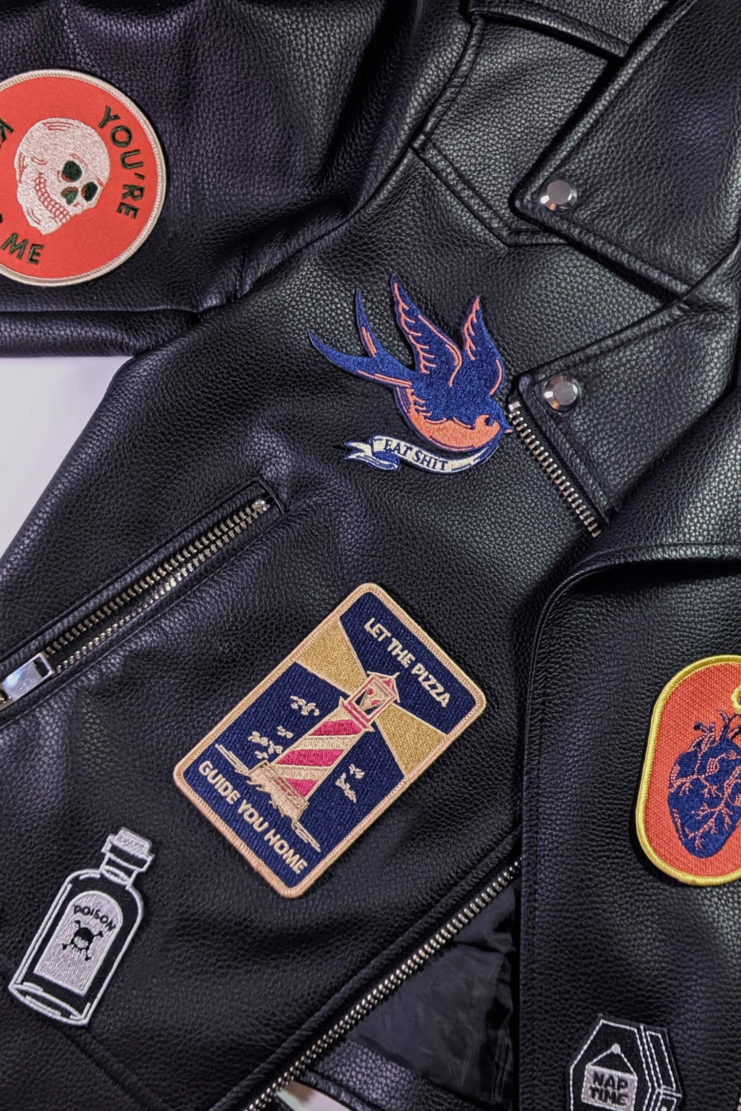 A vintage-style embroidered patch from Badaboöm Studio, 
featuring a flying bird in navy blue and orange, with a banner reading 'EAT SHIT' below, shown on black leather jacket with other patches. 