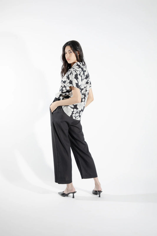 Back view of a woman wearing the Alisme Pants by Eve Lavoie, slightly cropped wide-legged pants with side patch pockets and an elastic waist. She is wearing them with a floral top and standing in front of a white background. 