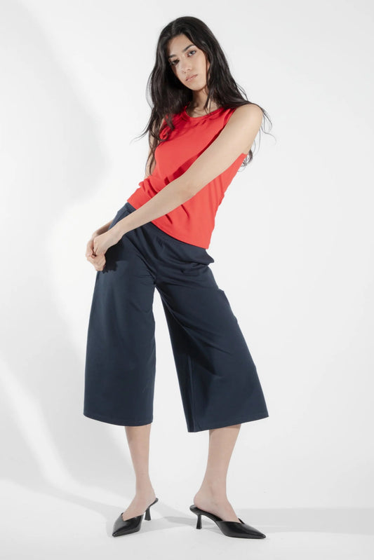 A woman wearing the Chara Pants by Eve Lavoie in Navy, featuring a wide pull-on waistband and wide legs that hit just below the knee. She is wearing them with a coral top and standing in front of a white background. 