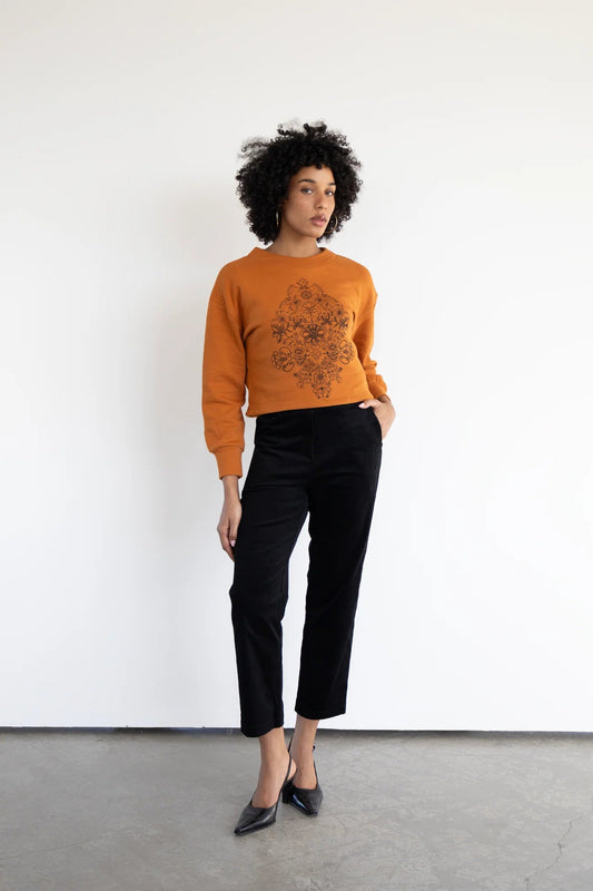 A woman wearing the Hanay Sweatshirt by Eve Lavoie in Burnt Orange, a slightly cropped sweatshirt with a hand-printed silkscreen design. she is wearing it with black pants and standing in front of a white background. 