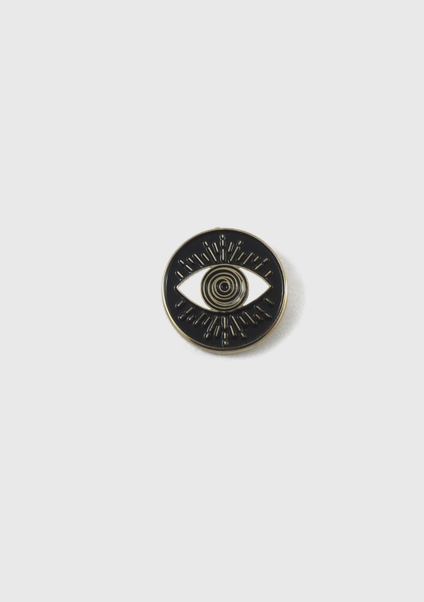 The Eyeball pin by Badaboöm Studio, a 
circular black enamel pin with a gold-tone metal border and an eye design in the centre, is shown against a white background.