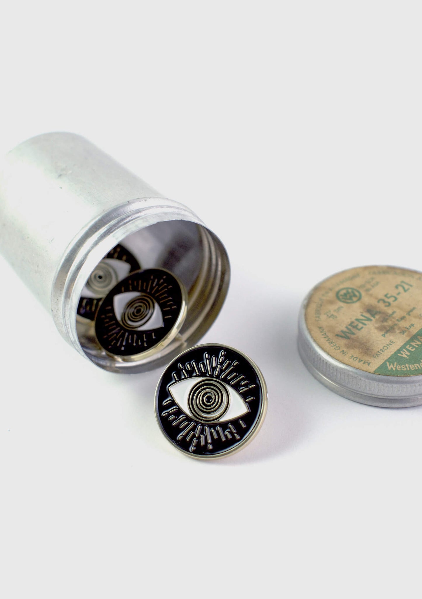 A number of The Eyeball pins by Badaboöm Studio, circular black enamel pins with a gold-tone metal border and an eye design in the centre, are shown spilling out of a silver container against  a white background.