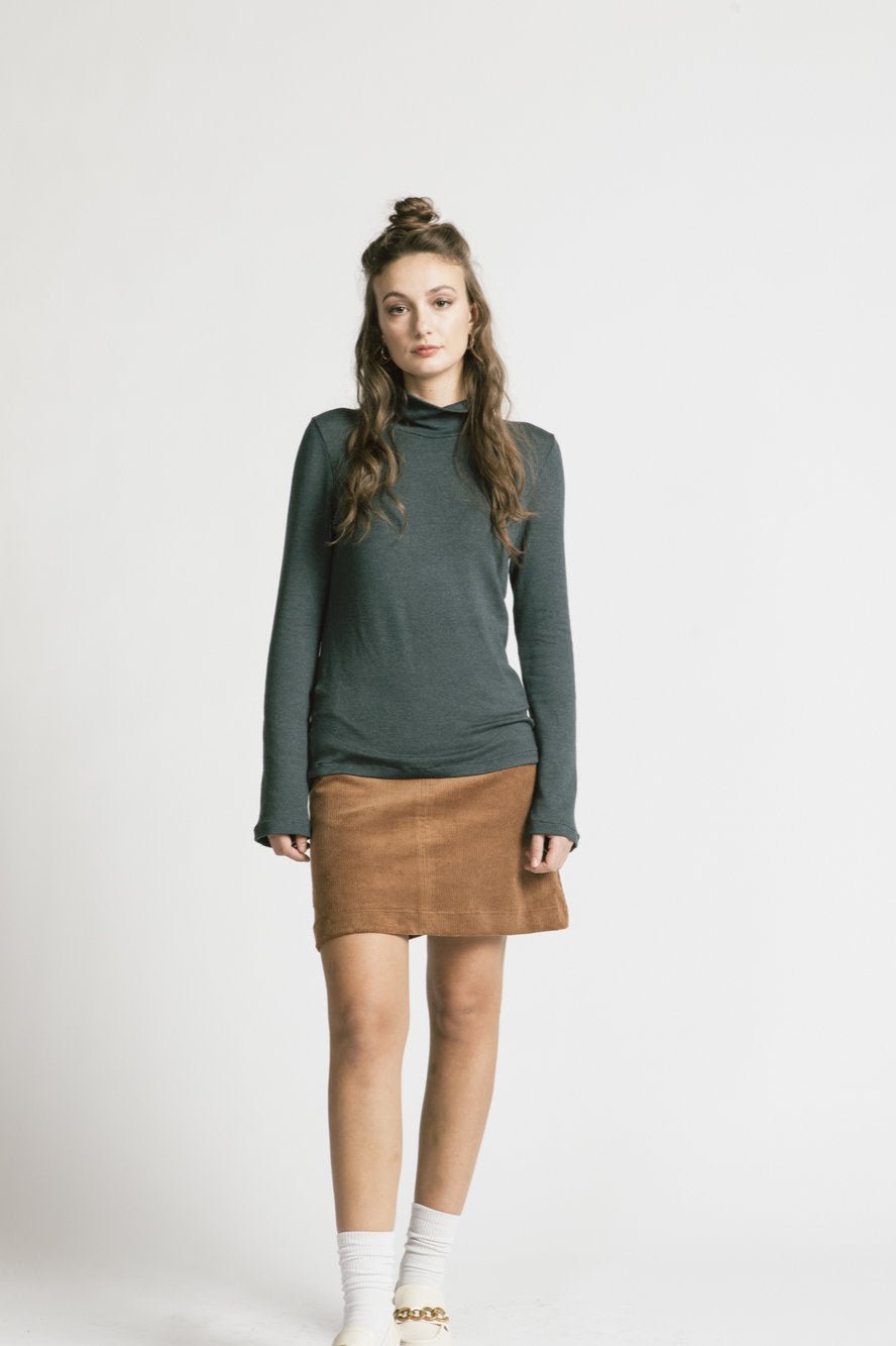 A woman wearing the Forever Turtleneck by Allison Wonderland in Denim, with a brown skirt, standing in front of a white background 