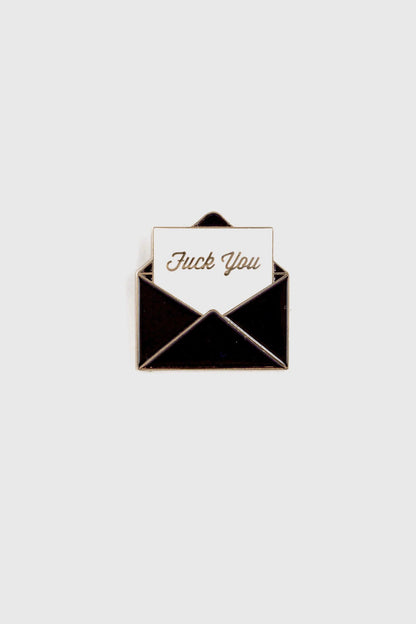 The Fuck You Pin by Badaboöm Studio, featuring a black envelope and white piece of paper that reads "fuck you" in gold lettering, is shown against a white background. 