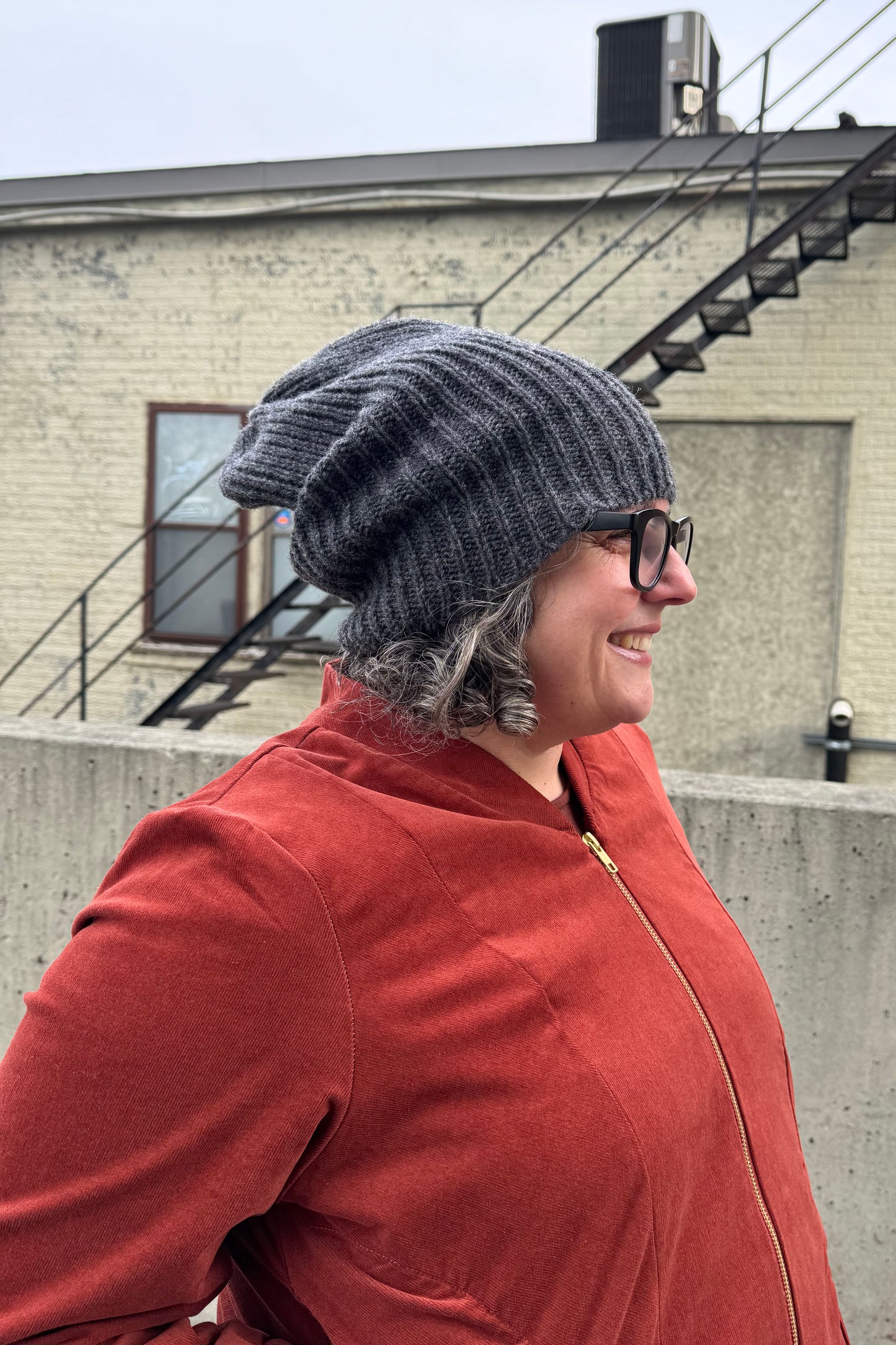 A woman wearing the Ribbed Beanie by Charlevoix Pure Laine in Charcoal. She is wearing it with the Redrawn Zip Jacket by Eve Lavoie and is standing outside. 