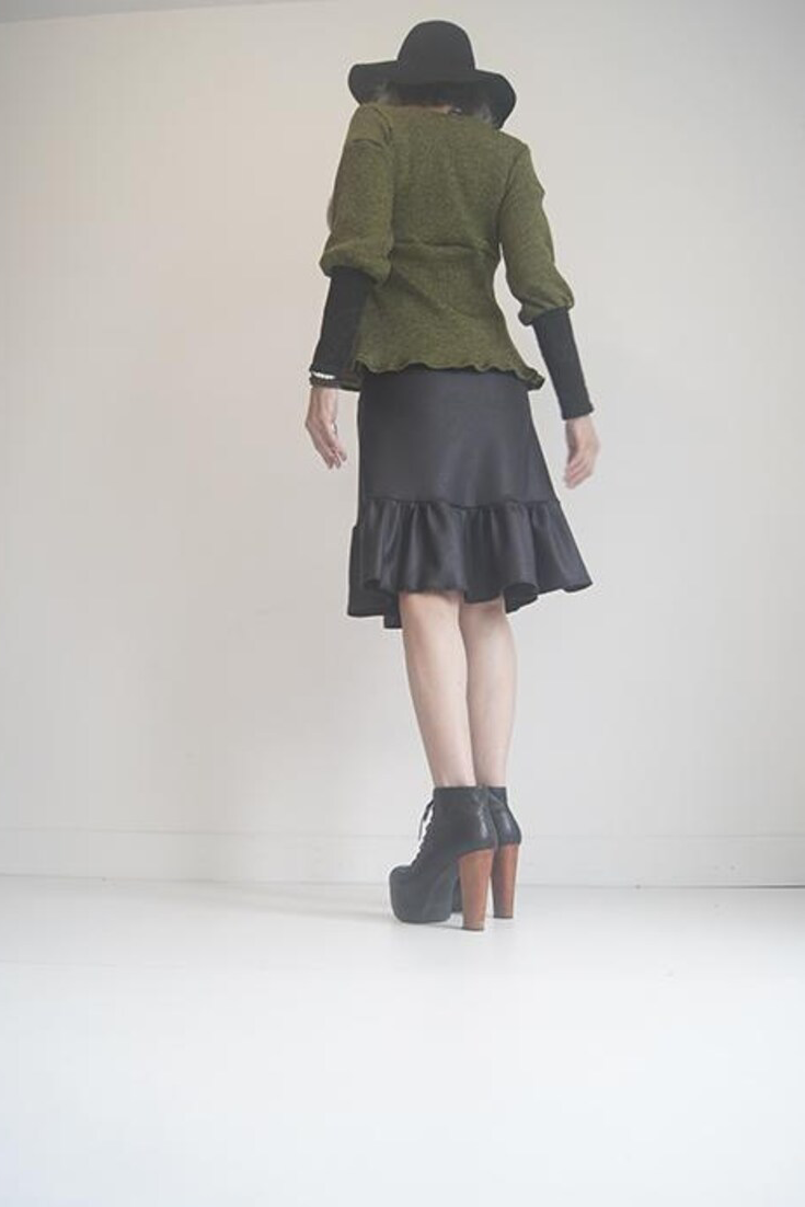 Back view of a woman wearing the Athena Top by SI Design in Olive Green, with a square neckline, and empire waist, and bishop style sleeves with long cuffs in black lace. She is wearing it with a black skirt and is standing in front of a white background 