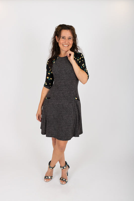 A woman wearing the Raphaelle Dress from Infime in Black, an A-line knee-length dress with 3/4 raglan sleeves that feature a floral print and pockets with a floral detail. She is standing in front of a white background