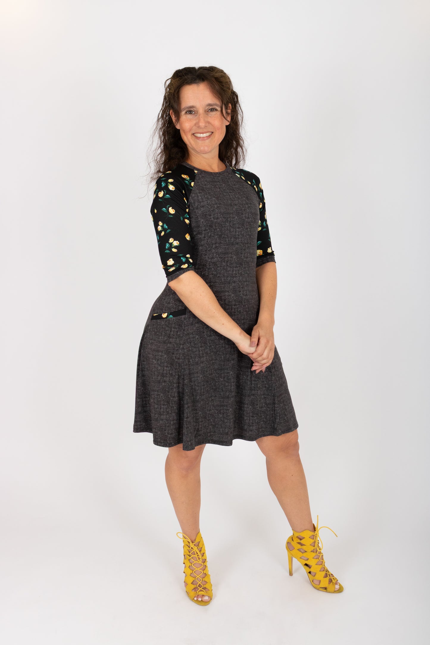 A woman wearing the Raphaelle Dress from Infime in Black, an A-line knee-length dress with 3/4 raglan sleeves that feature a floral print and pockets with a floral detail. She is standing in front of a white background