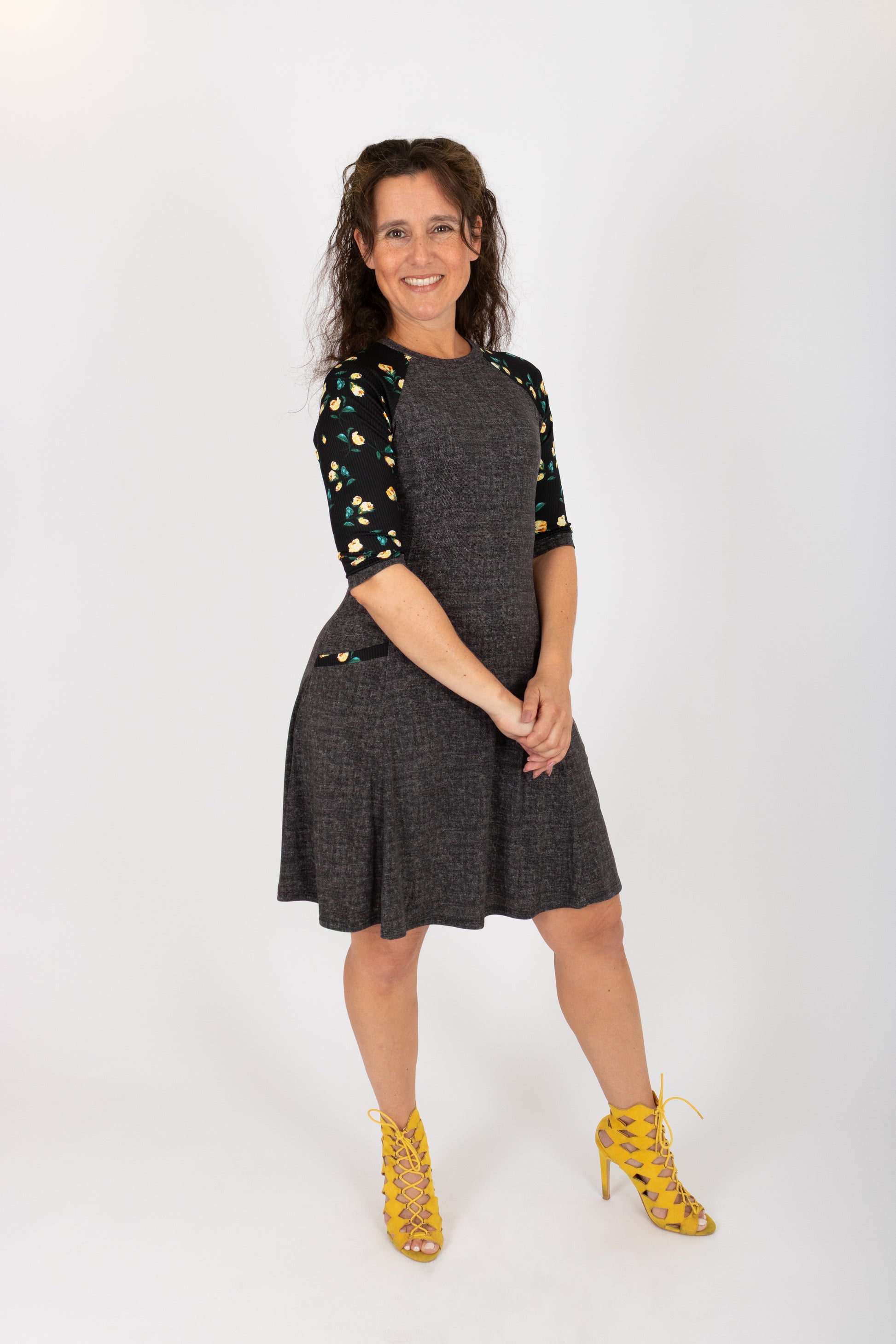 A woman wearing the Raphaelle Dress from Infime in Black, an A-line knee-length dress with 3/4 raglan sleeves that feature a floral print and pockets with a floral detail. She is standing in front of a white background