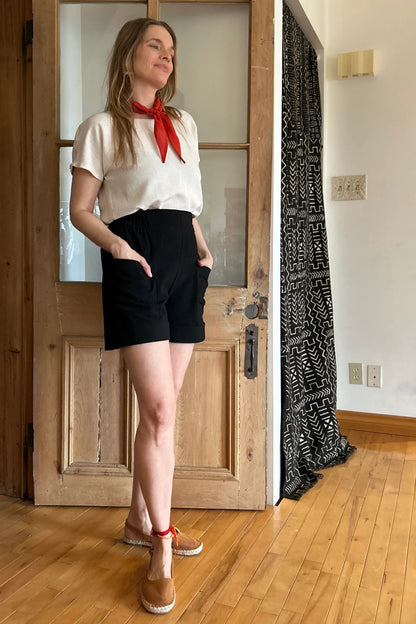 A woman wearing the Iris Shorts by Kazak in Black, with a smooth front waist, elastic at the back, large pockets, and mid-thigh length. She is wearing them with a cream top and red scarf and standing in a room with a door. 