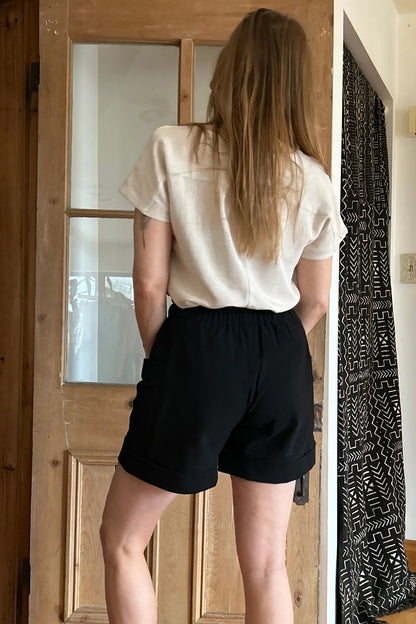 Back view of a woman wearing the Iris Shorts by Kazak in Black, with a smooth front waist, elastic at the back, large pockets, and mid-thigh length. She is wearing them with a cream top and red scarf and standing in a room with a door. 