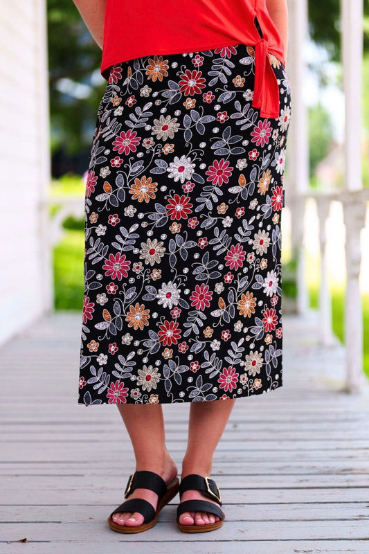 Waist down view of a woman wearing the Fiesta 7/8 Skirt by Rien ne se Perd in Colourful Daisies, a slightly A-line skirt with an elastic waist and side slits. She is wearing it with a red top and standing on a wooden porch.