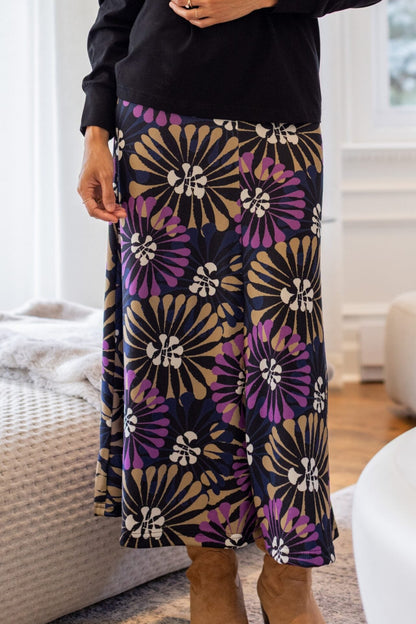 Waist down view of a woman standing in a white living room wearing the Hudson Skirt by Rien ne se Perd in Floral, a midi-length skirt with a bold purple, gold, and white floral burst pattern. She is wearing it with the Becancour Top in Black. 
