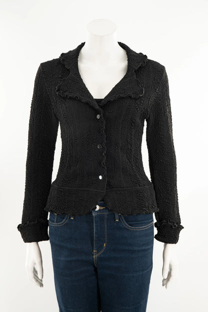 The Essential Jacket - Jasper by Karkass is shown on a mannequin in front of a white background. It's a black textured jacket with a ruffled collar, button closure, and frill details at the cuffs and hem. 