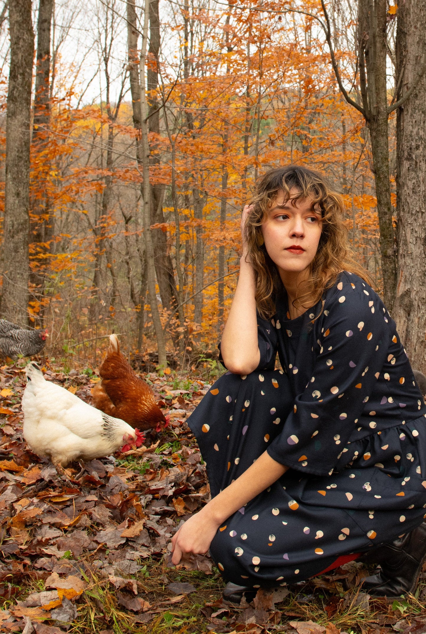 A woman wearing the Persea Dress by Kazak in Midnight Moon print, a below-the-knee length dress with 3/4 sleeves, a removable belt, and large patch pockets. She is crouching in a forest with chickens beside her. 