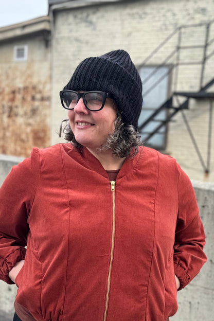 A woman wearing the Ribbed Beanie by Charlevoix Pure Laine in Black. She is wearing it with the Redrawn Zip Jacket by Eve Lavoie and is standing outside. 