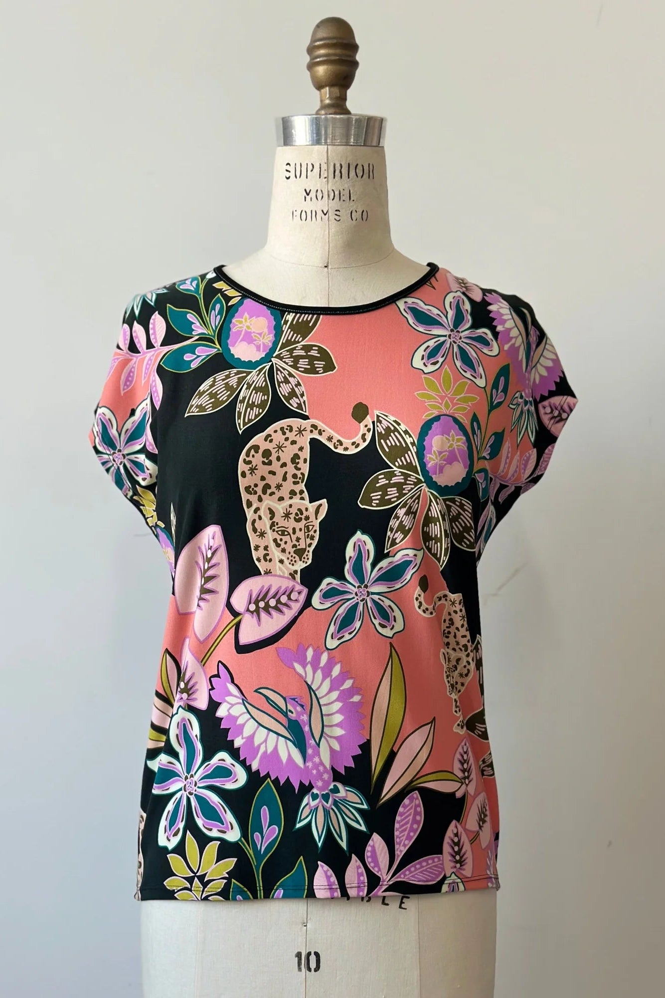 The Badia T-shirt by Kollontai in Bubblegum, featuring a patterned front with leopards, flowers and birds and a solid black back with cap sleeves and a hi-low hem, is shown on a mannequin in front of a white background.