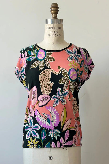 The Badia T-shirt by Kollontai in Bubblegum, featuring a patterned front with leopards, flowers and birds and a solid black back with cap sleeves and a hi-low hem, is shown on a mannequin in front of a white background.