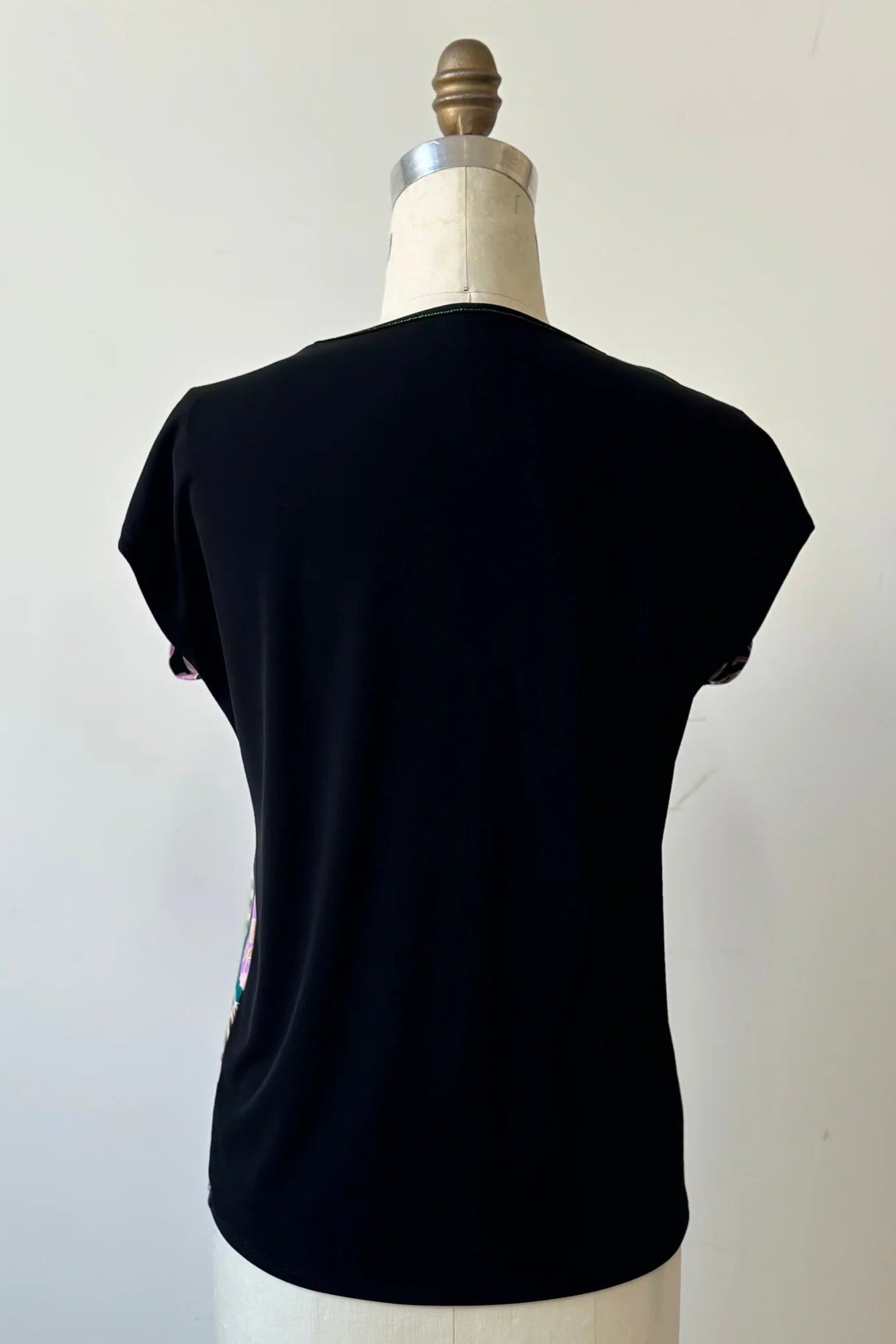 A back view of the Badia T-shirt by Kollontai in Bubblegum, featuring a patterned front with leopards, flowers and birds and a solid black back with cap sleeves and a hi-low hem, is shown on a mannequin in front of a white background.